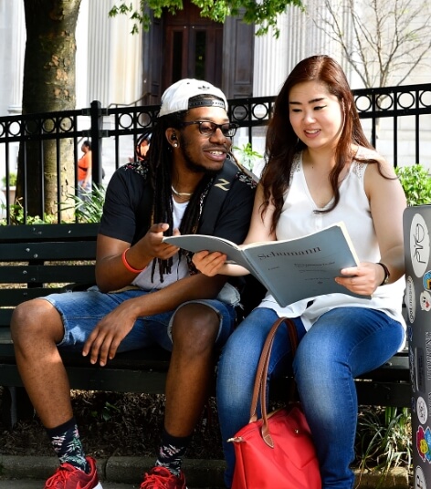 Two students sit outside, on a bench, reading from the same booklet.