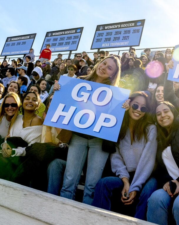 Student with a sign reading Go Hop stands in the front row of a crowded stadium.